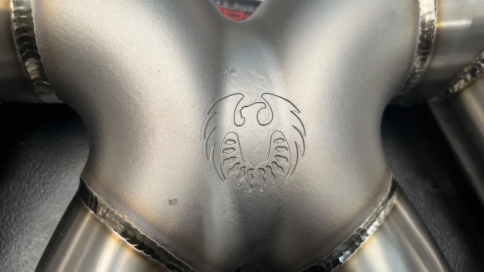Close-up of a custom stainless steel exhaust component with a laser-etched phoenix logo and visible welds.