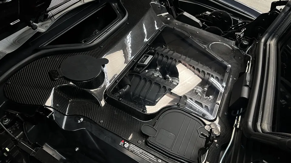 A detailed view of a supercar engine bay featuring carbon fiber components and a clear engine cover showcasing the intake manifold.
