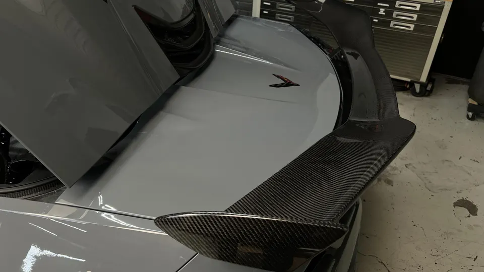 A grey Corvette C8 with a carbon fiber rear wing installed in a shop setting, showcasing the aerodynamic upgrade.