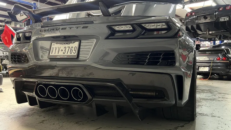 Rear view of a gray Corvette C8 with a large carbon fiber wing, aftermarket exhaust, and diffuser in a performance shop.