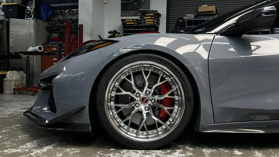 A grey Corvette C8 with custom chrome wheels, red brake calipers, and carbon fiber aero parts in a shop.