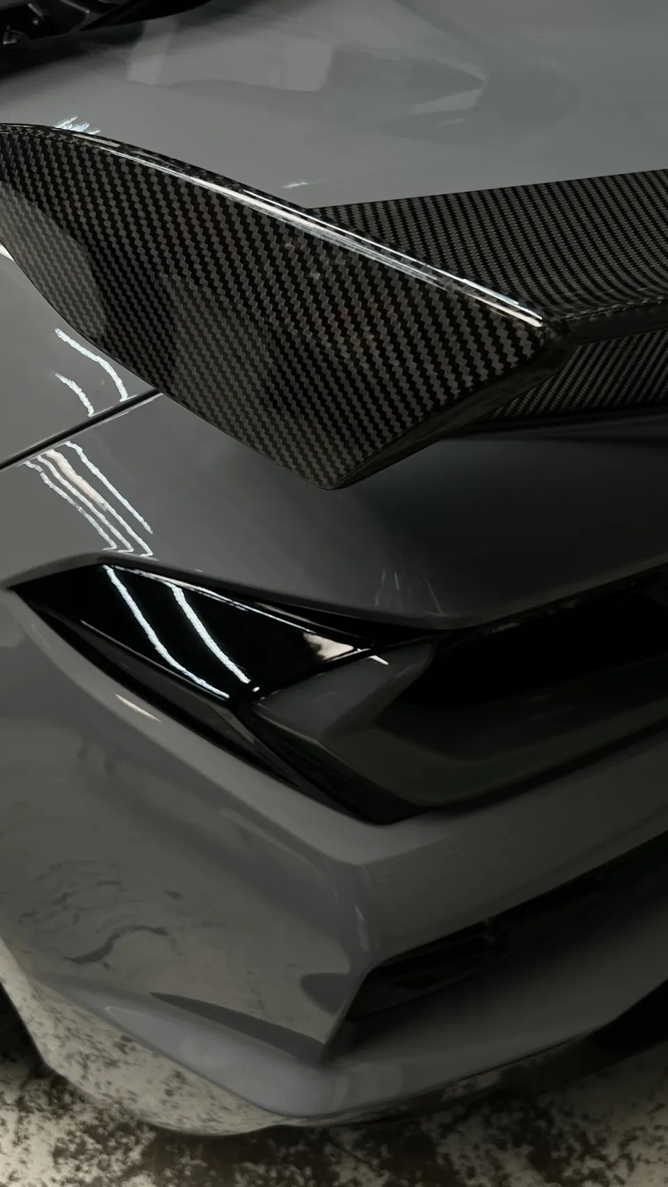 Close-up of a grey car's front bumper featuring a carbon fiber aerodynamic element.