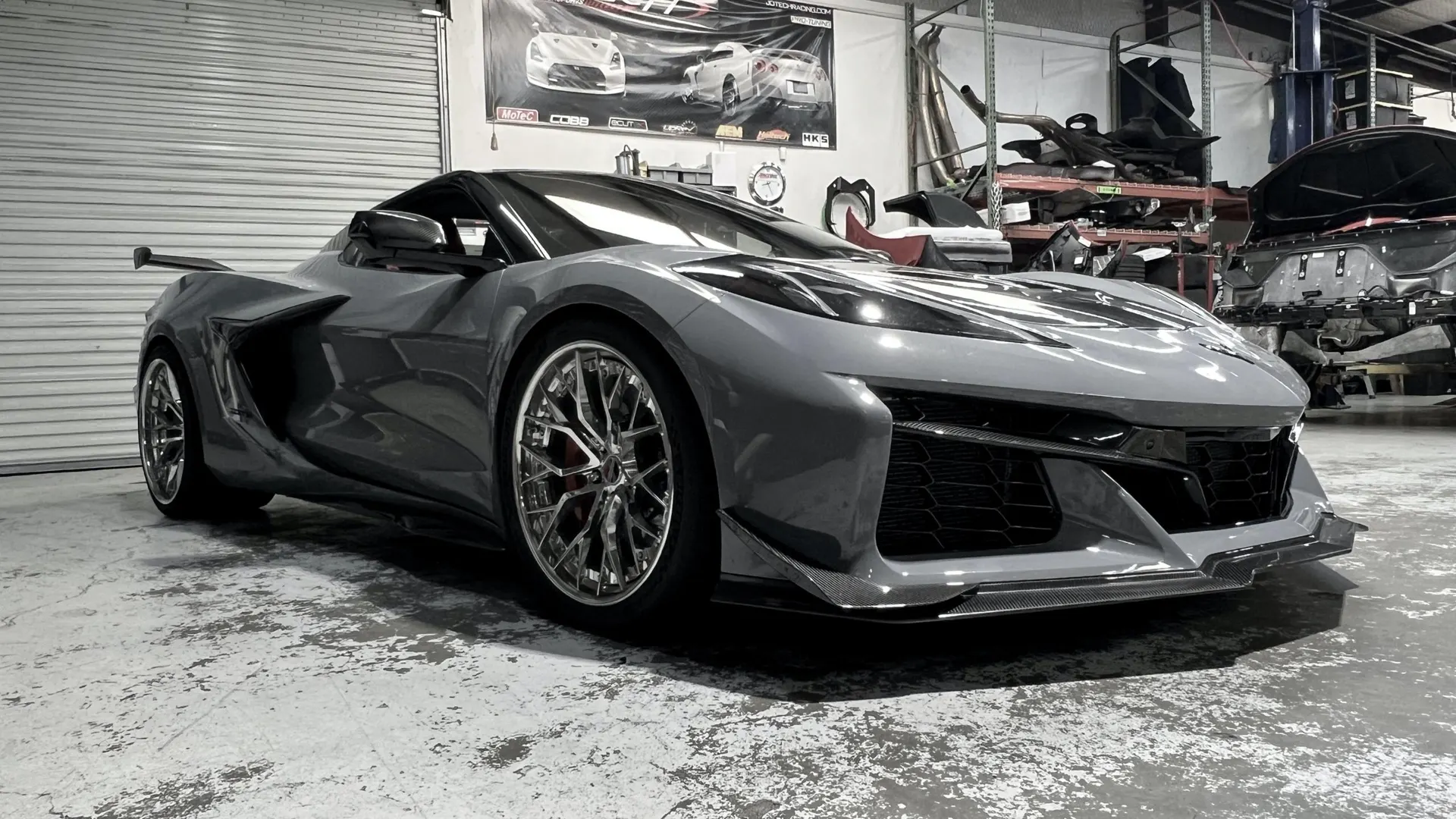 A grey Chevrolet Corvette C8 with aftermarket wheels and carbon fiber aero parked in a performance tuning shop.