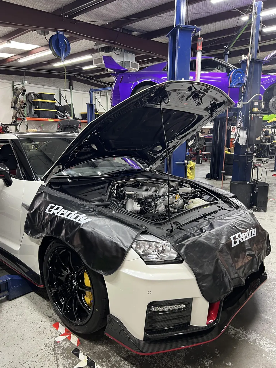 A white Nissan GT-R with its hood open, protected by GReddy fender covers, in a performance tuning shop.