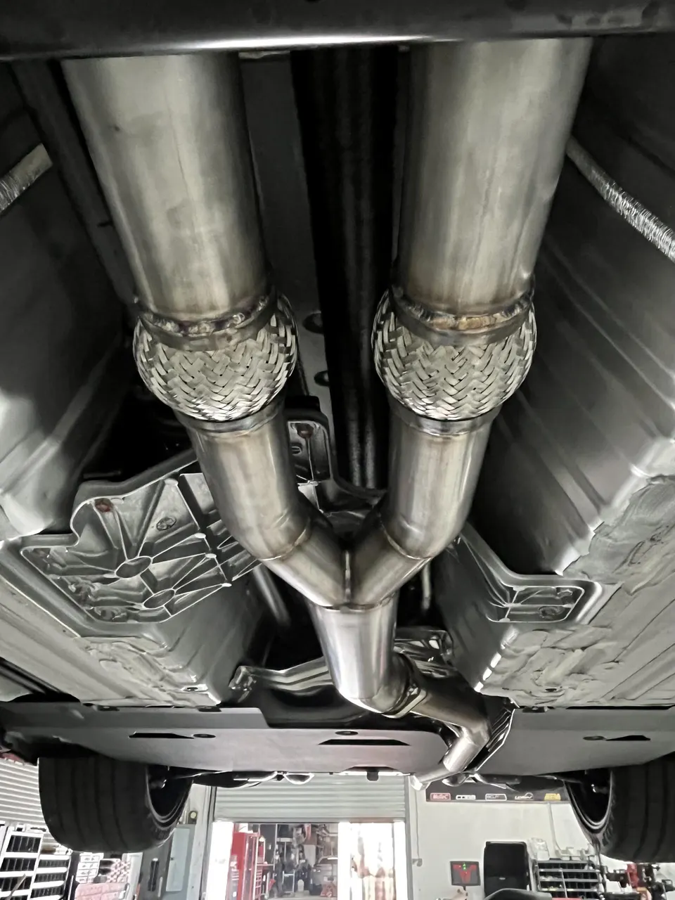 Underside view of a performance car on a lift, showcasing a custom stainless steel exhaust system with flexible pipes.