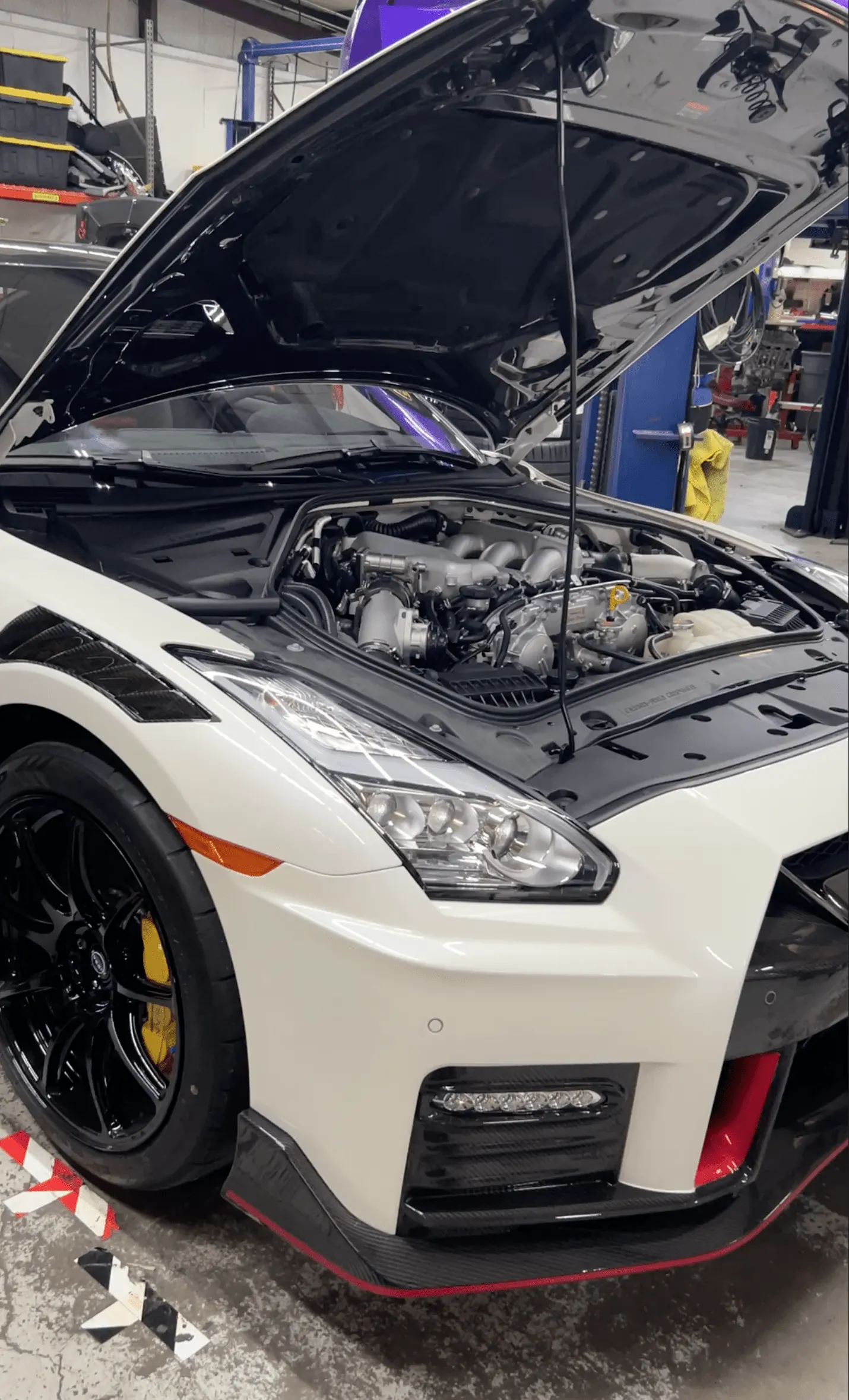 A white Nissan GT-R with its hood open, revealing the engine bay and intake manifold in a performance shop.