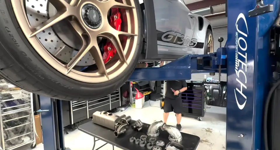 A silver Porsche GT3RS on a blue car lift in a Jotech Motorsports shop, with exhaust components on a table below.