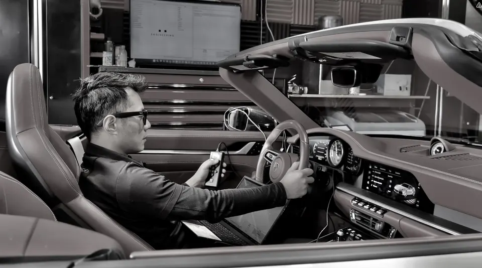 A person in a Porsche convertible interior using a laptop and handheld device for performance tuning in a shop.