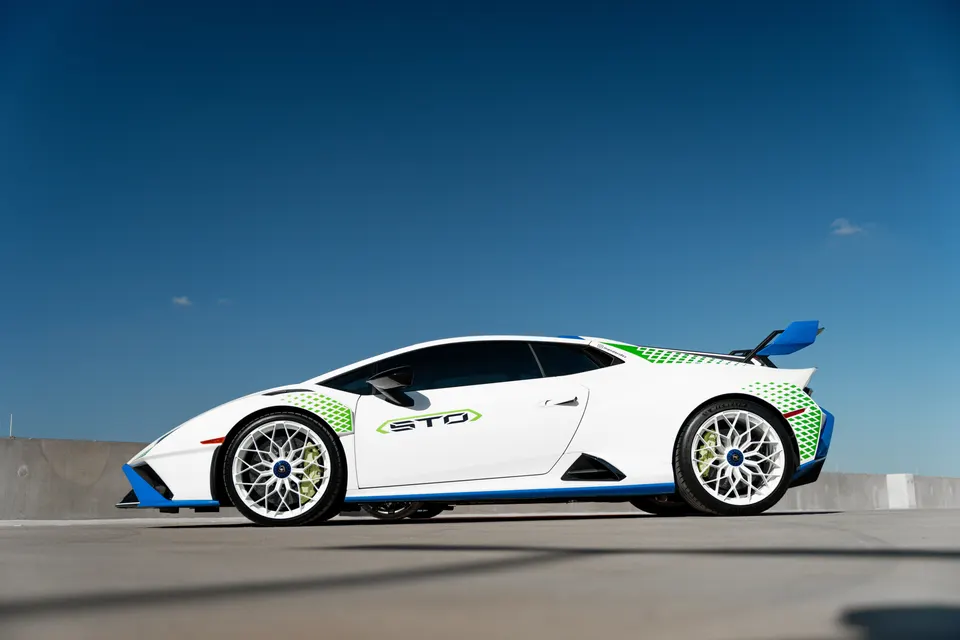 A white Lamborghini Huracan STO with blue and green accents and custom white wheels parked outdoors under a blue sky.