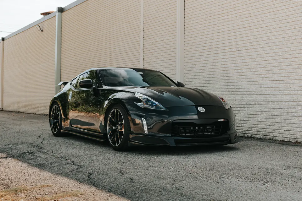 A black Nissan 370Z sports car with aftermarket wheels parked on an asphalt road next to a light brick wall.