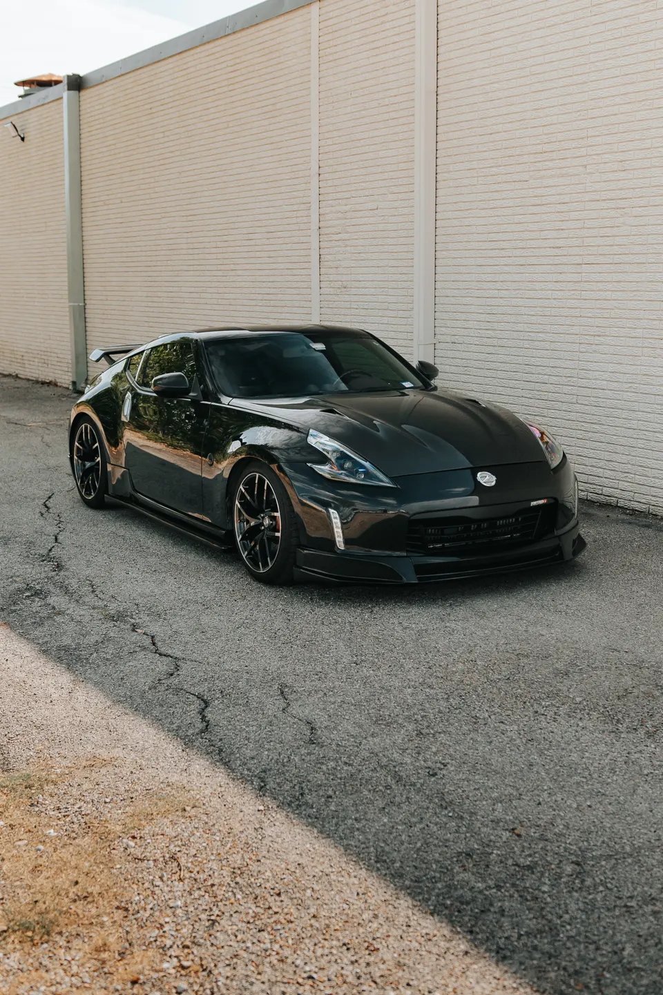 Black Nissan 370Z with aftermarket aero kit and wheels parked on an asphalt road next to a brick wall.