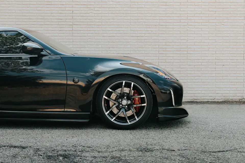 Dark grey Nissan 370Z with custom multi-spoke wheels, red brake calipers, and carbon fiber side skirts parked by a brick wall.
