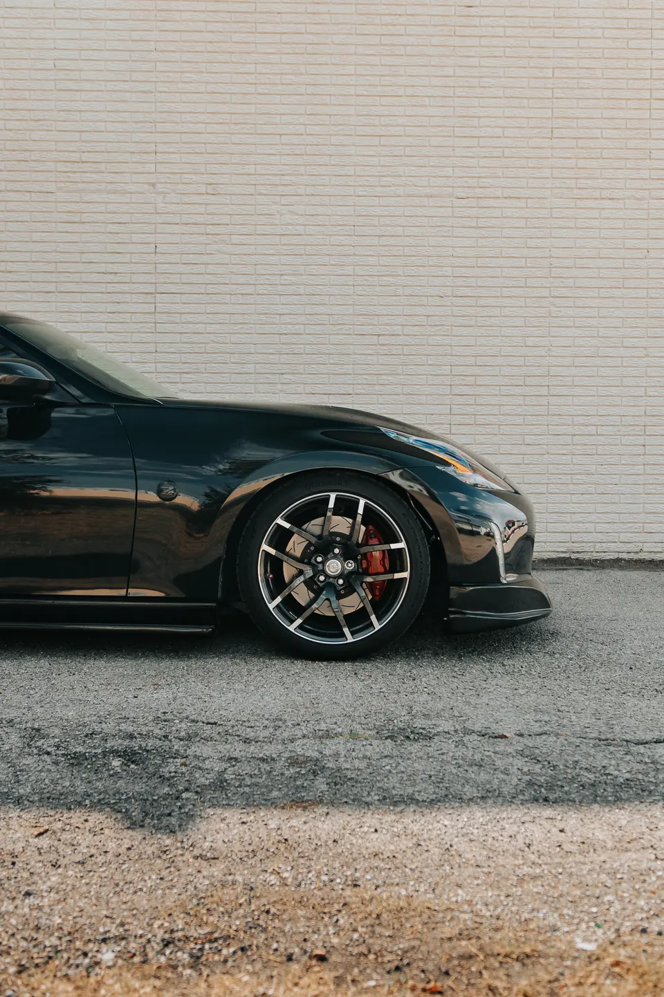 A black Nissan 370Z with custom multi-spoke wheels and red brake calipers parked against a white brick wall.