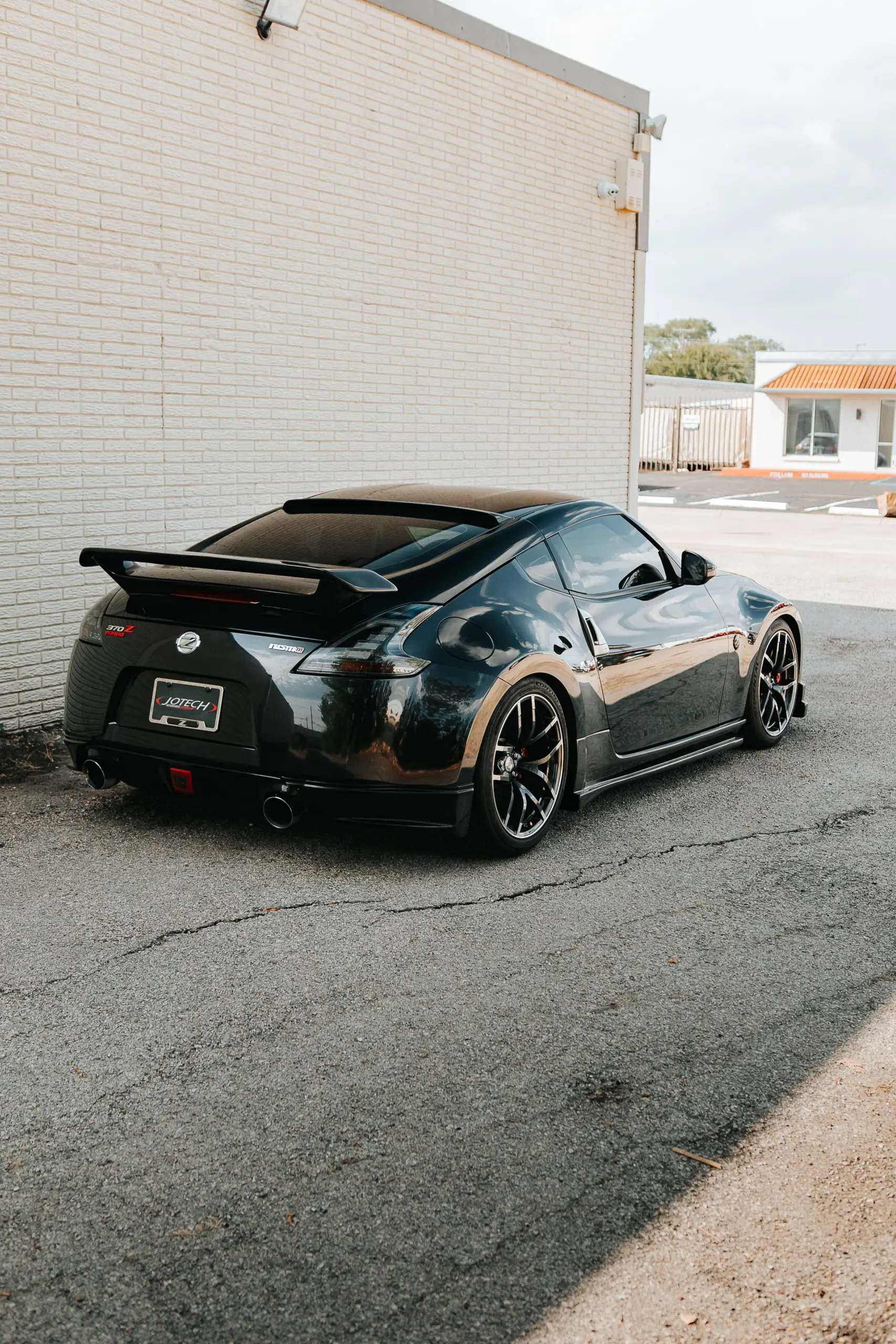 Black Nissan 370Z with aftermarket wheels, rear spoiler, and dual exhaust tips parked next to a white brick wall.