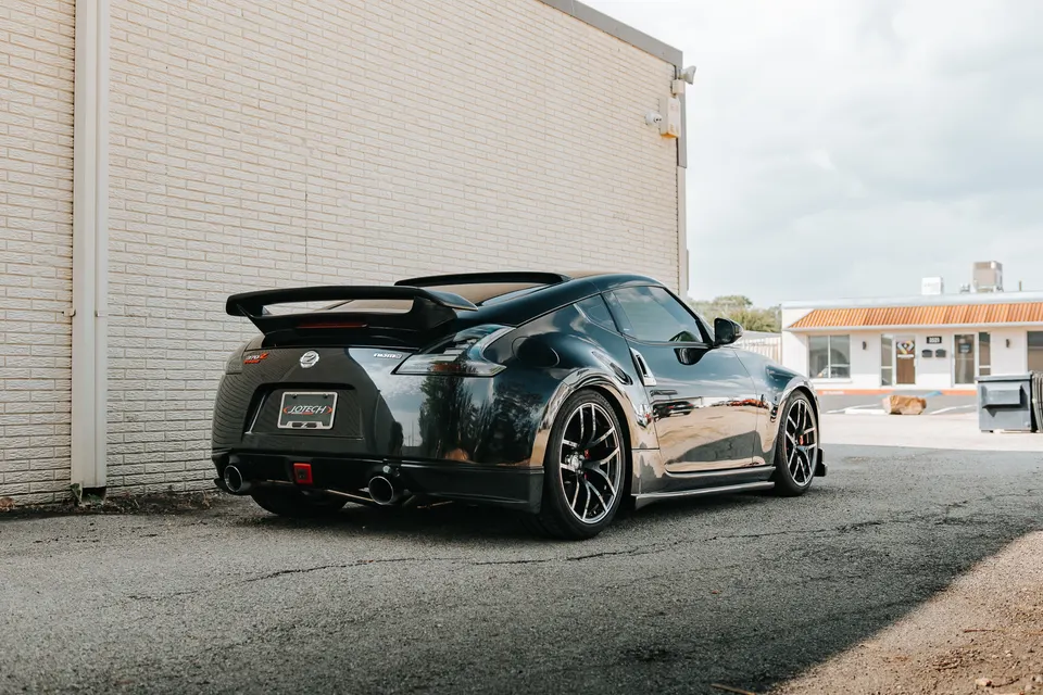 A black Nissan 370Z with an aftermarket wing and custom exhaust parked outside a building on a sunny day.