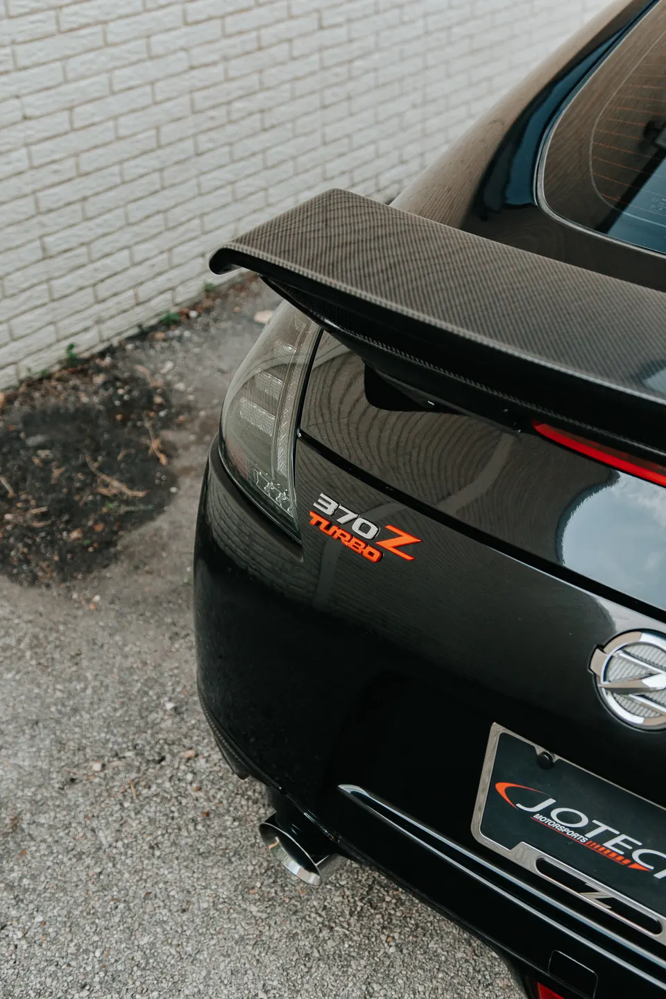 Rear view of a black Nissan 370Z with a carbon fiber spoiler, '370Z TURBO' badge, and Jotech Motorsports license plate frame.