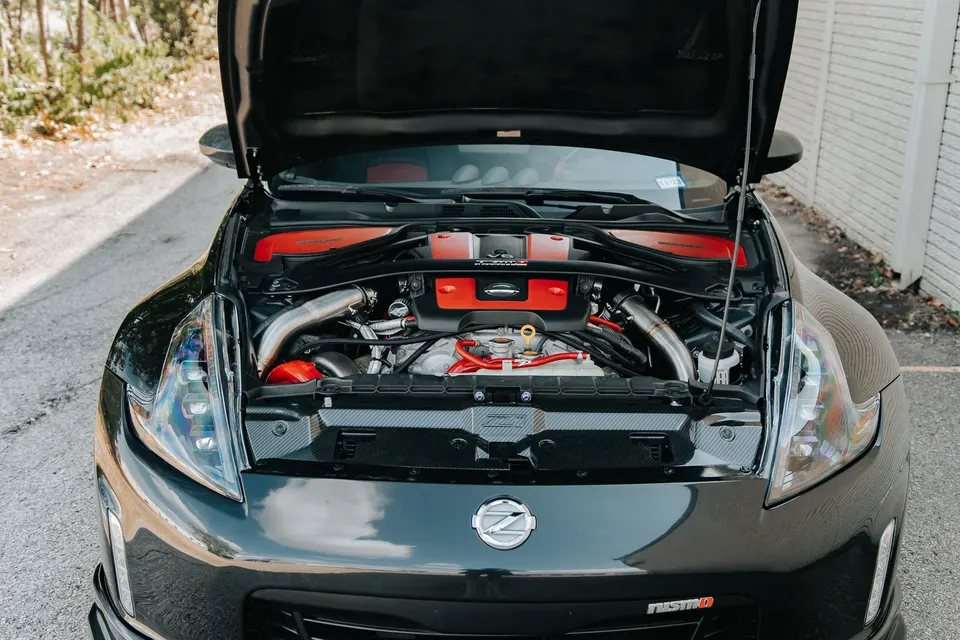 Front view of a black Nissan 370Z Nismo with the hood open, showcasing a custom engine bay with red accents and polished induction pipes.