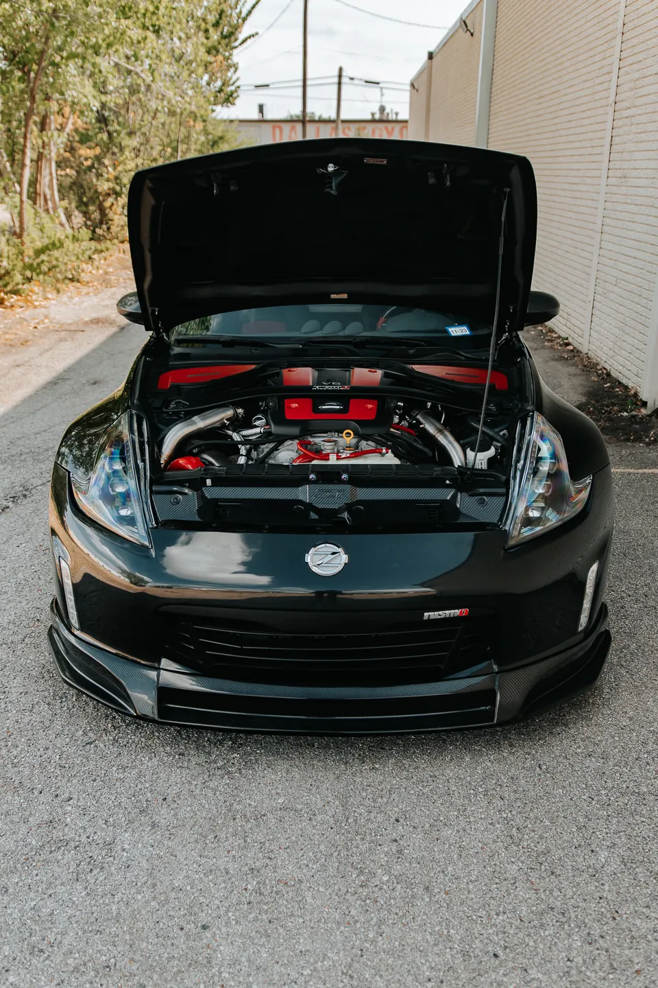 A black Nissan 370Z Nismo with its hood open, showcasing a custom engine bay with red accents and aftermarket induction.