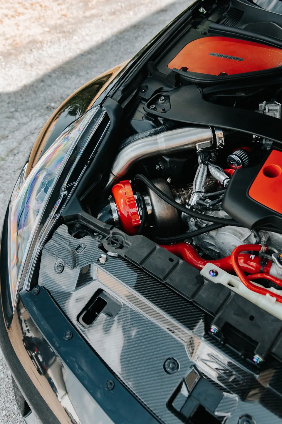 A close-up view of a performance car's engine bay featuring a red turbocharger, custom piping, and carbon fiber trim.
