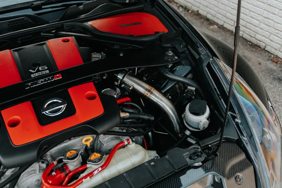 A close-up view of a Nissan VQ VVEL engine bay with a Nismo engine cover and aftermarket intake pipe.
