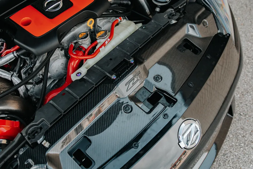 Close-up of a Nissan Z engine bay featuring a carbon fiber radiator shroud, red silicone hoses, and a custom engine cover.