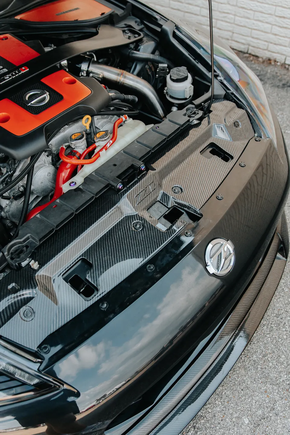 A close-up of a Nissan 370Z engine bay featuring a red engine cover, aftermarket intake, and carbon fiber trim.