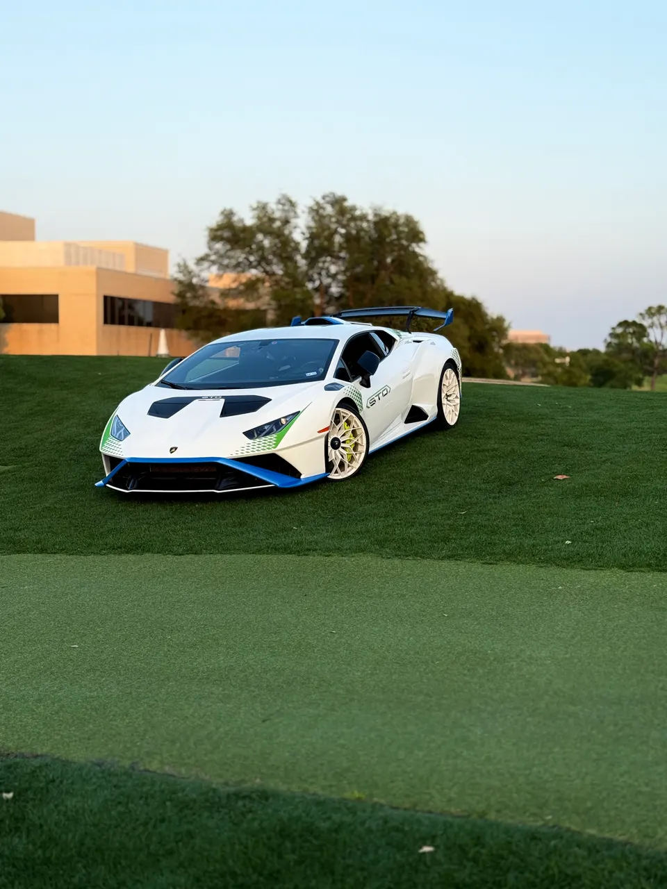 A white Lamborghini Huracan STO with blue and green accents parked on a grassy hill with buildings in the background.