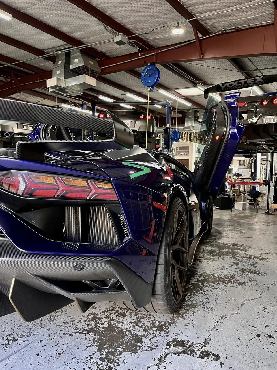 A purple Lamborghini Aventador with a large carbon fiber wing and diffuser in a performance tuning shop.
