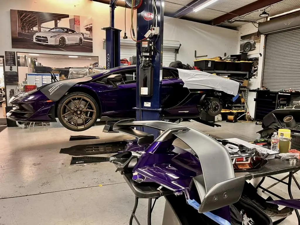 A purple Lamborghini Huracan on a lift in a performance shop, with a carbon fiber rear wing on a table.