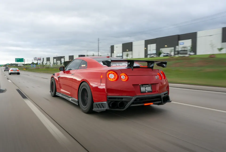 A red Nissan GT-R with a widebody kit and large aftermarket wing drives on a highway under a cloudy sky.