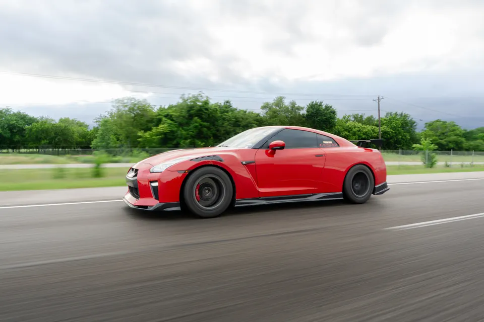 A red Nissan GT-R with aftermarket wheels and aero parts driving on a road under a cloudy sky.