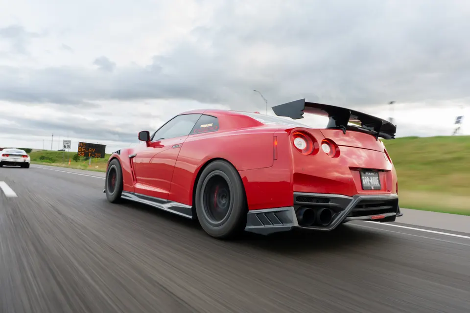 Red Nissan GT-R with an aftermarket wing and diffuser driving on a highway under a cloudy sky.