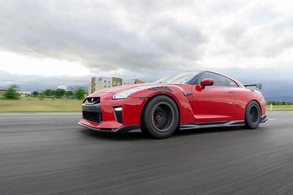 A red Nissan GT-R with aftermarket wheels and aero drives on a road under a cloudy sky, captured in a dynamic roller shot.