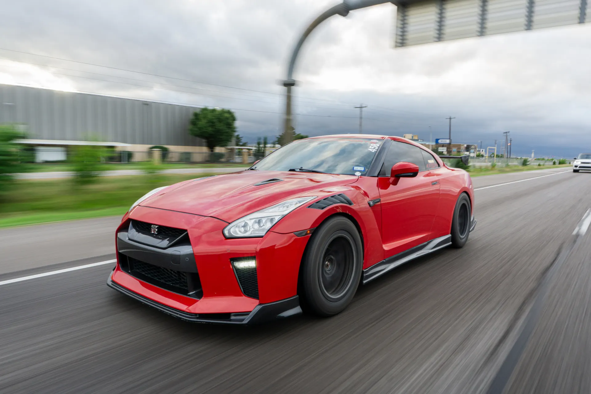 A red Nissan GT-R with an aftermarket aero kit and wheels driving on a road under a cloudy sky.