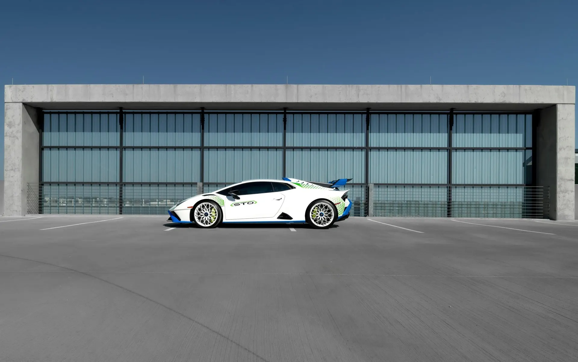 A white Lamborghini Huracan STO with blue and green accents parked in a modern concrete parking structure.