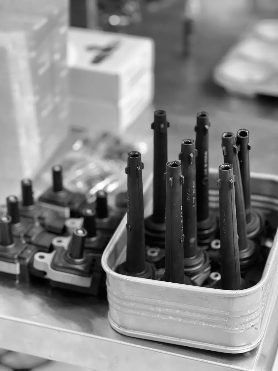 Black and white close-up of new ignition coils for a performance car engine on a metal workbench in a shop.