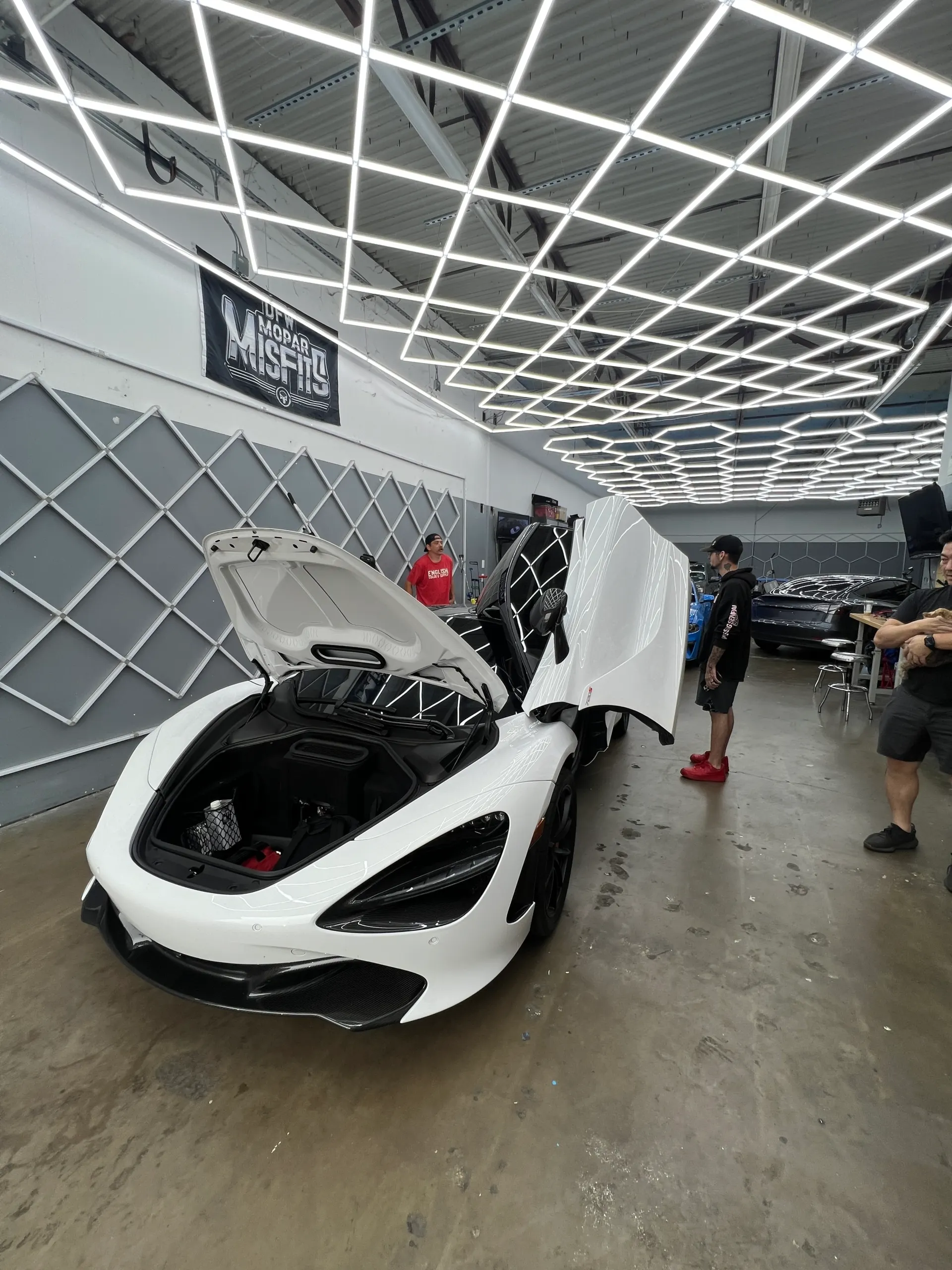 A white McLaren 720S with its frunk and butterfly doors open inside a detailing shop with distinctive grid lighting.