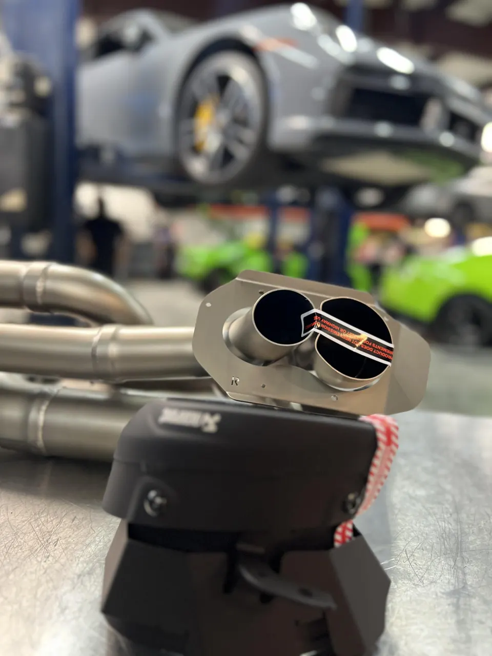 Close-up of an Akrapovic exhaust system with a Porsche on a lift in the background at a performance shop.