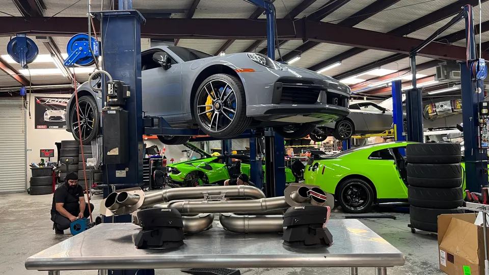 A grey Porsche 911 on a lift in a performance tuning shop, with an Akrapovic exhaust system on a table in the foreground.