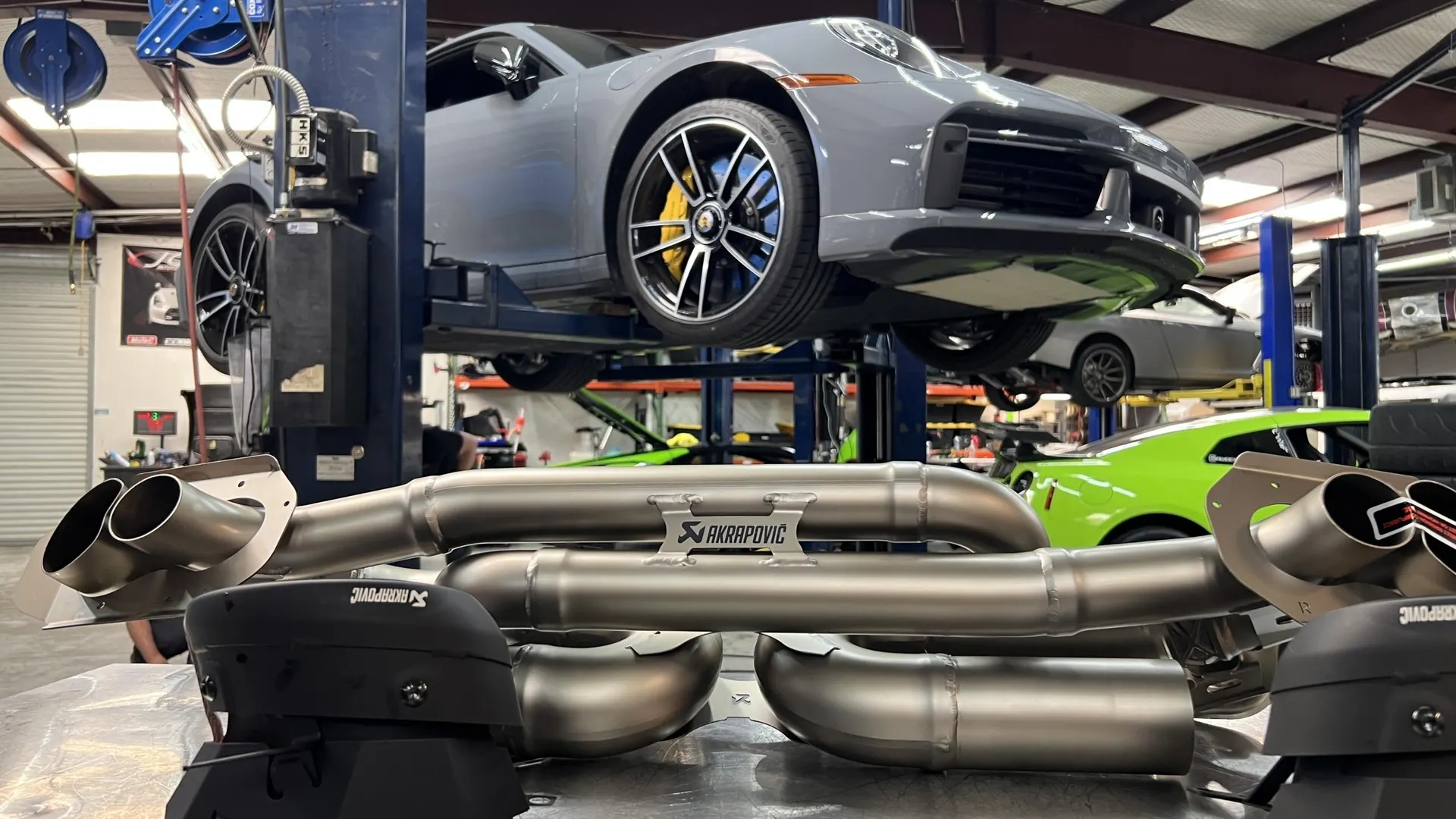 A grey Porsche 911 on a lift in a performance shop with an Akrapovic exhaust system on the floor, ready for install.