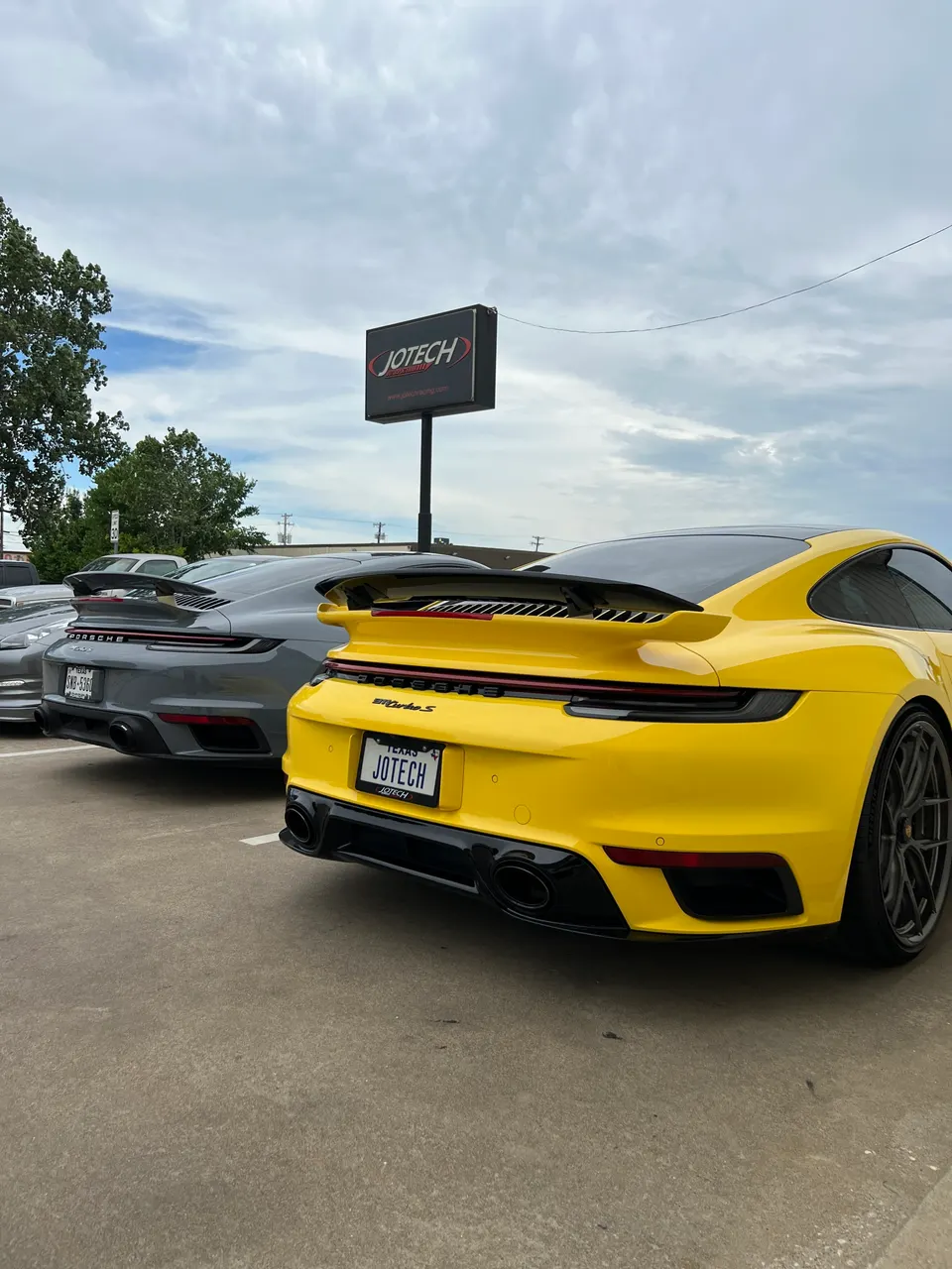 Yellow and grey Porsche 911 Turbo S cars parked outside Jotech Motorsports, showcasing aftermarket exhaust and rear wing.