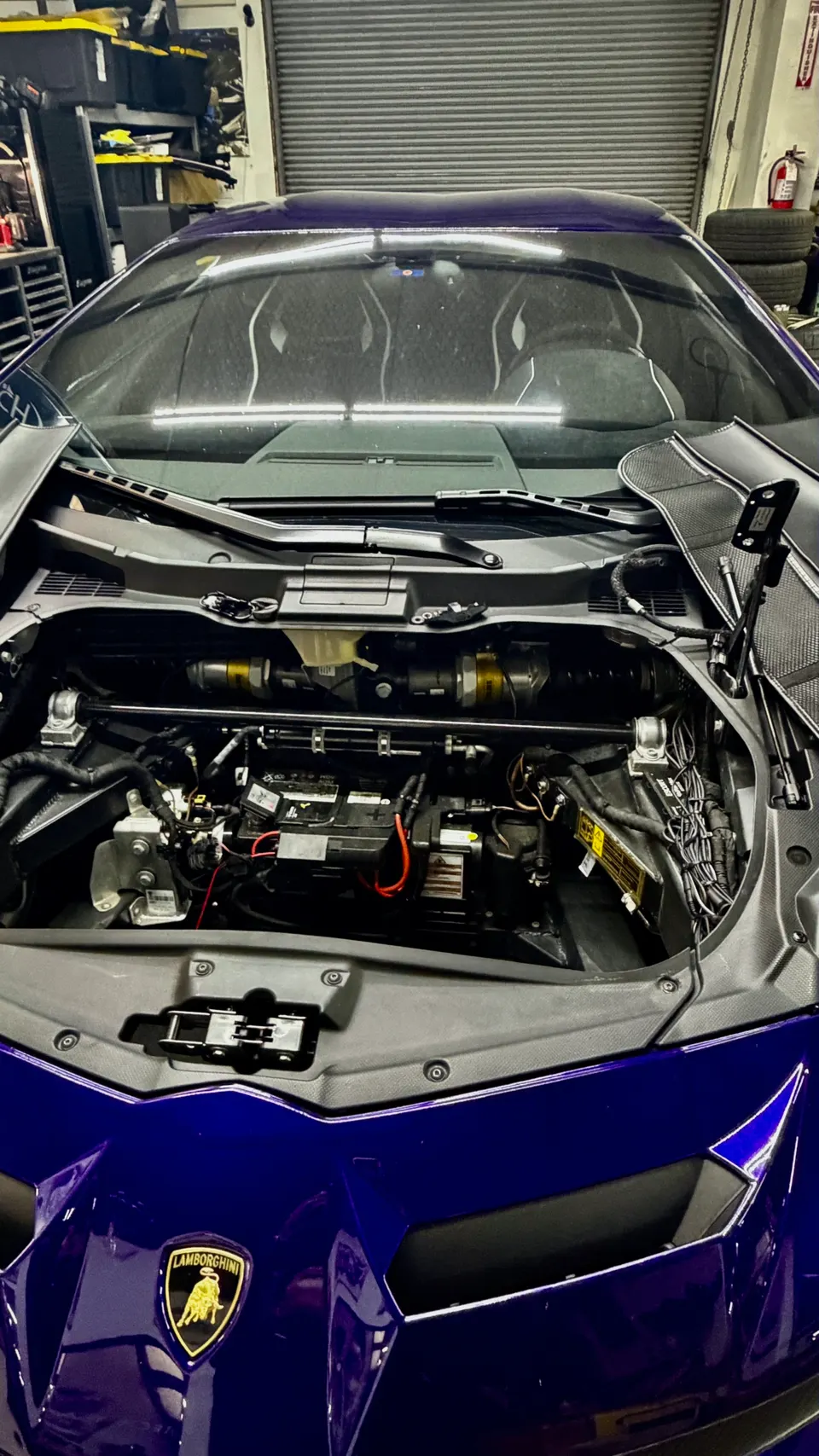 Purple Lamborghini with its front trunk open, revealing the battery and wiring in a performance tuning shop.
