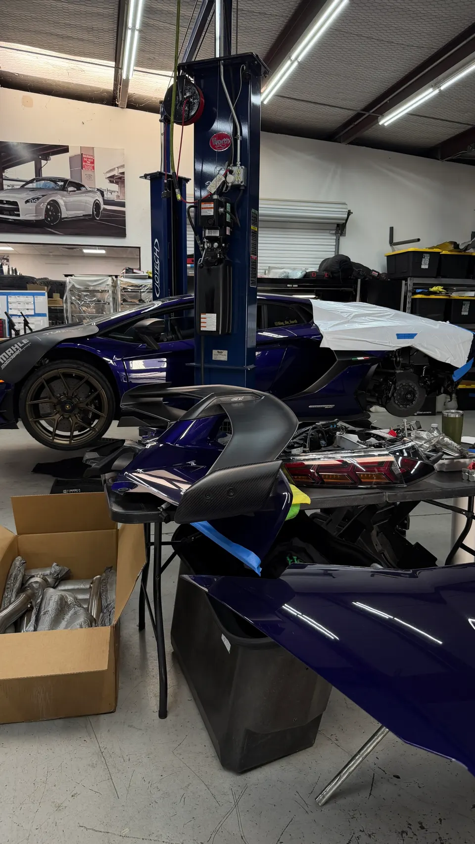 A purple Lamborghini Aventador on a car lift in a Jotech Motorsports shop, with a carbon fiber wing and exhaust parts on display.