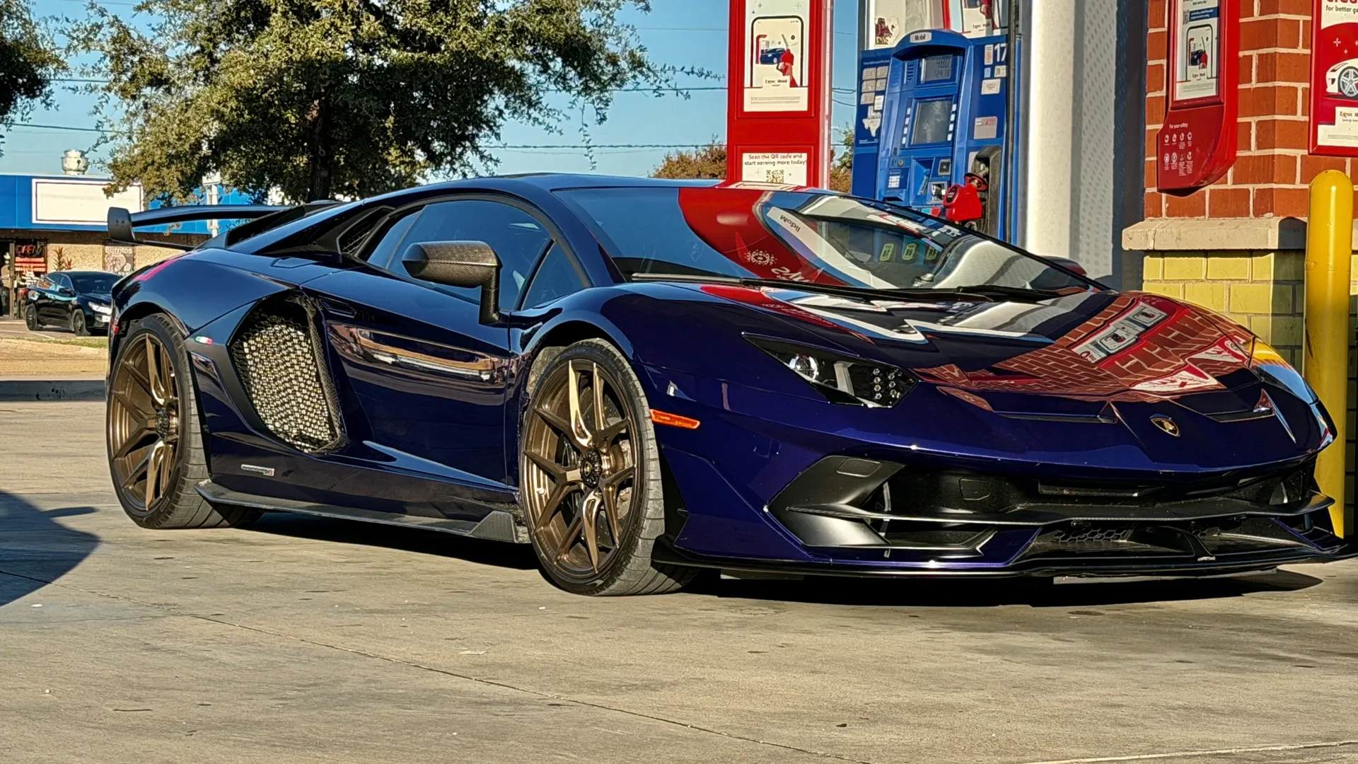 A dark blue Lamborghini Aventador with bronze wheels parked at a gas station on a sunny day.