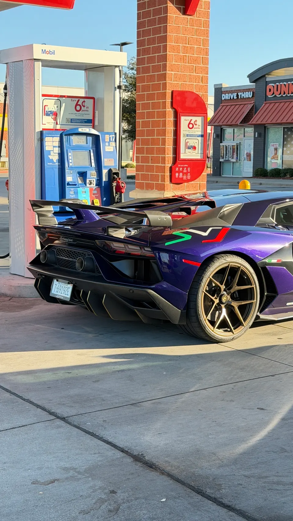 Purple Lamborghini Aventador SVJ with a large rear wing and bronze wheels at a gas station.