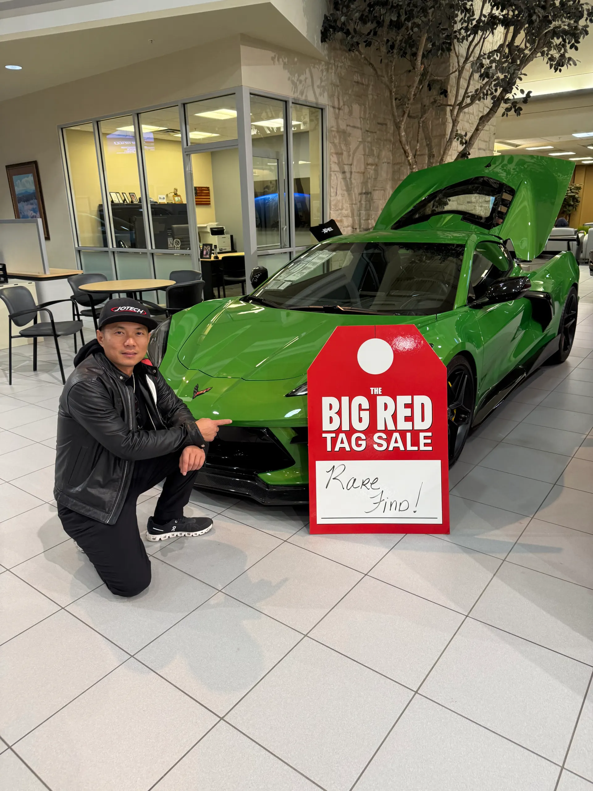 Man kneeling next to a green Chevrolet Corvette C8.R with its engine bay open at a dealership during a big red tag sale.