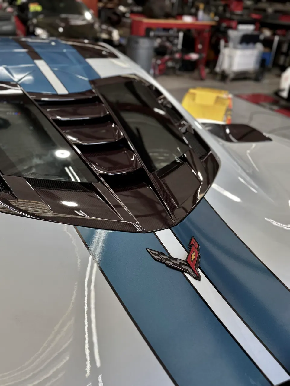 Close-up of a white Chevrolet Corvette C8 hood with blue racing stripes and a carbon fiber vent in a performance shop.