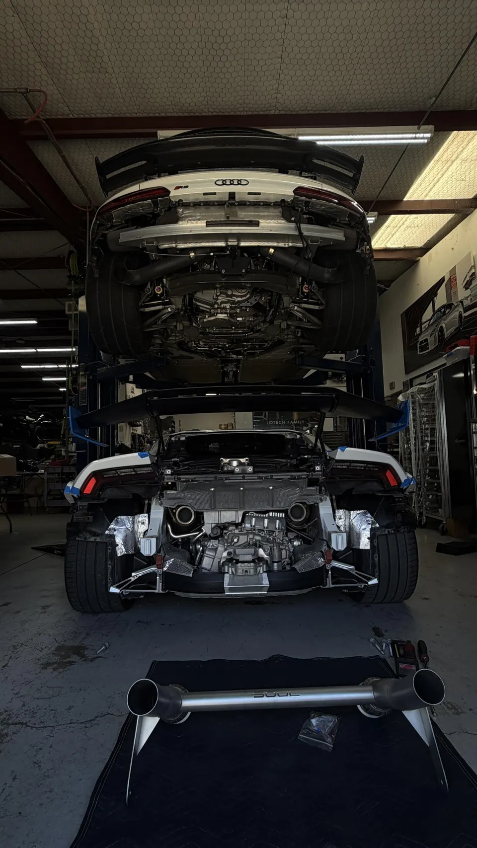 Two sports cars, an Audi R8 and a Lamborghini Huracan, on lifts in a performance tuning shop with an aftermarket exhaust on the floor.
