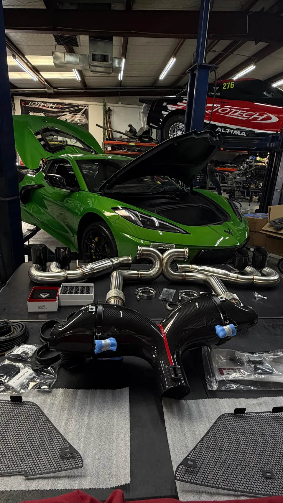 A green Corvette C8 on a lift in a Jotech Motorsports shop with a performance exhaust system and carbon fiber intakes on a table.