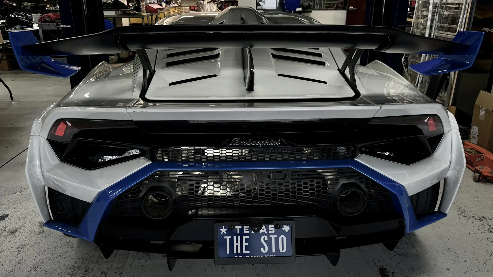 A white Lamborghini Huracan with a large black and blue rear wing and blue rear diffuser in a shop.